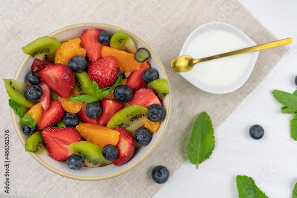 Fototapeta Fresh, healthy chopped fruit salad in a bowl on white  background. Served with yoghurt. Top view. Strawberry, blueberry, kiwi, orange, mint..