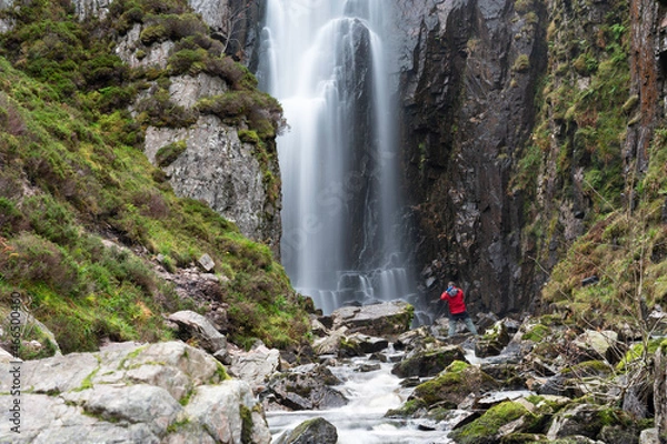 Fototapeta Photographer at Wailing Widows Falls, a famous waterfall in Assynt, North West Highlands of Scotland. On NC500 route. Smooth flow of water, stream, rocks, cliff edges either side with greenery.