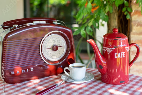 Obraz A white cup with coffee stands near a coffee pot and a radio in a retro style on a table in a summer garden. Outdoor breakfast theme.