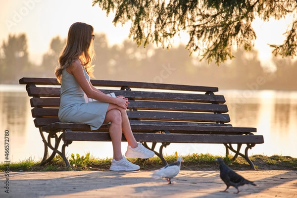 Fototapeta Lonely woman sitting on lake side bench enjoying warm summer evening. Solitude and relaxation concept