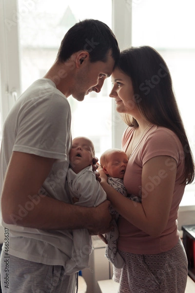 Fototapeta Loving family with newborn twins. Yong parents standing face to face near the window. Cute and tender pfoto of twins sleeping in mother's and father's arms. Siluette photo. Selective focus. Film grain