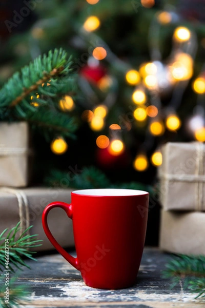 Fototapeta Coffee in a red cup on the rustic table with wrapped gifts, Christmas tree lights and spruce branches in the background