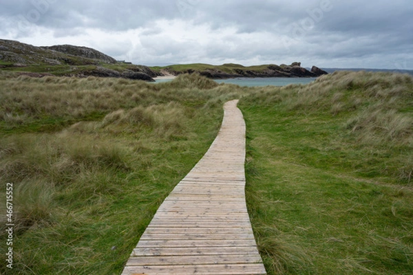 Fototapeta Clachtoll beach in Assynt, Scottish Highlands on a cloudy day with dramatic sky. Wooden boardwalk leading through grass to beach, sea and rocks visible, no people.