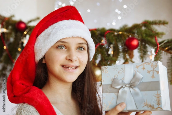 Obraz a smiling girl in a Christmas hat holds a gift box in her hands