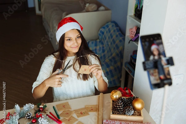 Obraz a smiling blogger girl in a Christmas hat makes an advent calendar and takes pictures on her phone