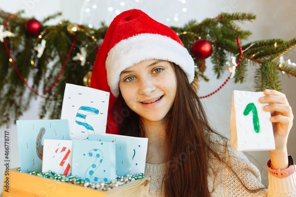 Obraz a happy girl in a Christmas hat holds an Advent calendar in her hands diy