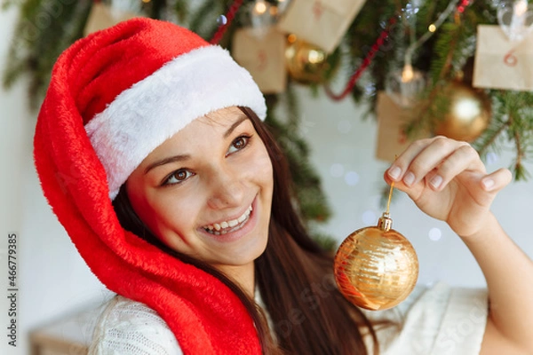 Obraz happy girl in a Christmas hat with a Christmas tree toy in her hand close-up