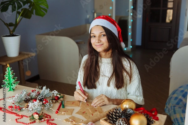 Obraz a smiling girl in a Christmas hat makes an advent calendar