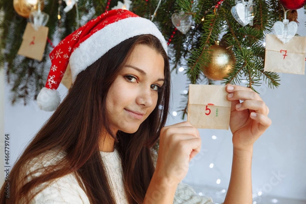 Obraz a smiling girl in a Christmas hat with an Advent calendar diy in her hands near the Christmas tree