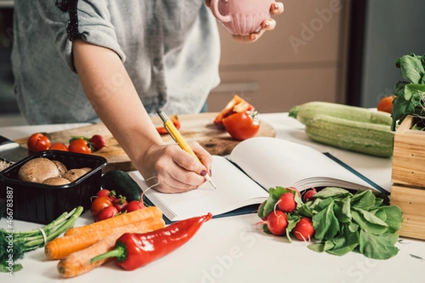 Fototapeta Close Up Photo of Woman Hand Writing Recipe in a Notebook and Holding a Cup of Coffee while Standing at Table Full with Fresh Colorful Vegetables for Homemade Lunch