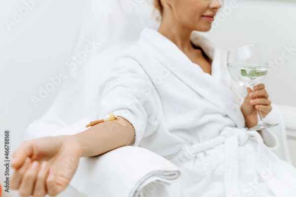Obraz Close up of calm female patient sitting with tube and needle during IV infusion. Girl holding glass with lemon water and relaxing