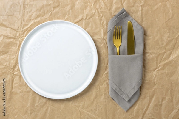 Fototapeta Cutlery, gold fork and knife in folded cloth napkin near white round empty plate on crumpled craft paper, mock up, flat lay, copy space. Table setting concept, environmentally friendly, zero waste