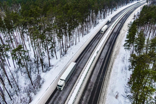 Fototapeta white trucks on the higthway. convoy  whith cargo driving by road seen from the air. Top view landscape. shooting from a drone. Cargo delivery in winter. Aerial view of snowy road in winter forest