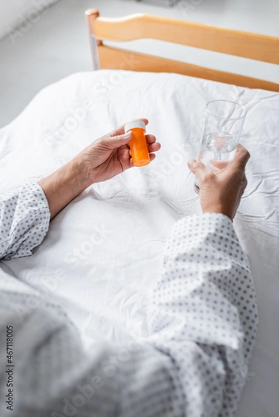 Fototapeta Cropped view of senior patient holding water and pills on bed in clinic