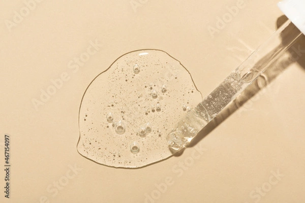 Obraz Spilled clear serum with bubbles and glass pipette on beige surface close upper view. Skincare and dermatological procedures