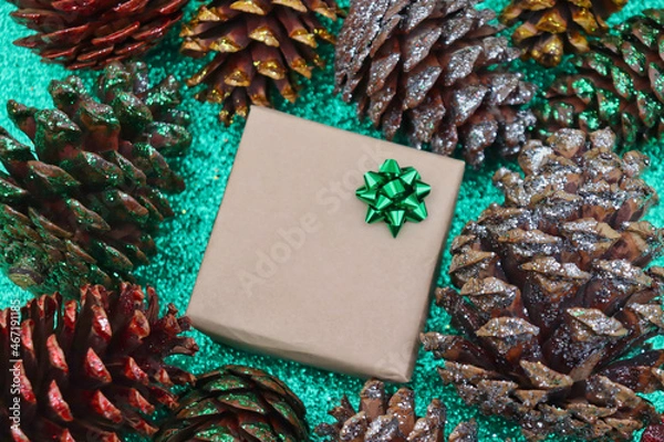 Fototapeta Present wrapped in brown paper with green bow surrounded by silver, green, red, and gold glitter pine cones on green glitter background.