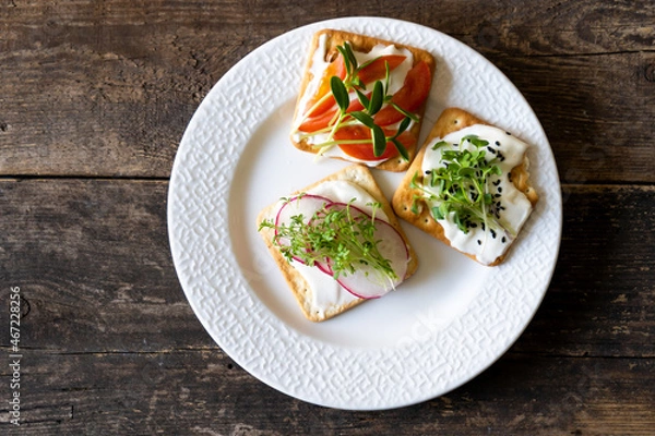 Obraz Set of Bruschett with cream cheese and tomato spread. Radish, micro green and flaxseed in food. Simple food on the plate. Breakfast in a white plate top view. Wooden background. Cracker