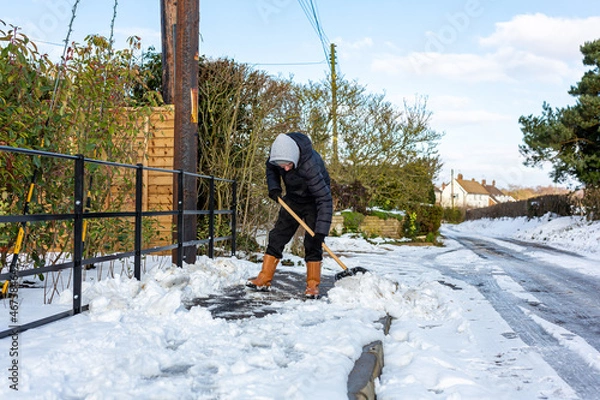 Fototapeta A young man clearing the footpath of snow and ice to make it safe to walk on during a heavy snowfall. Winter safety, clearing snow and ice concept