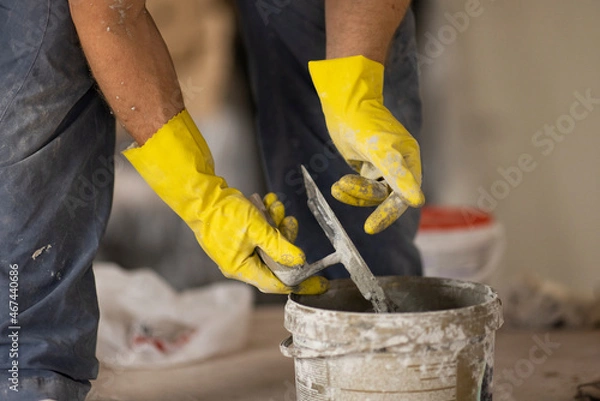 Fototapeta Hold a spatula in your hands and dilute the plaster in a bucket. The hands are wearing yellow gloves. The builder makes repairs in the apartment.