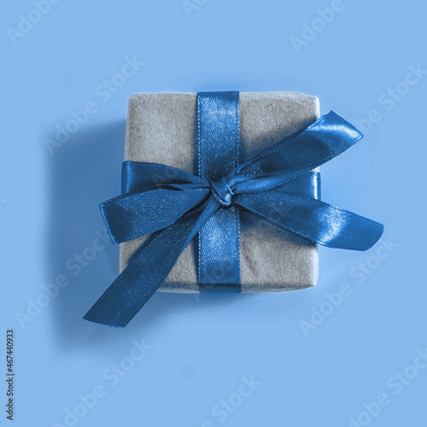 Fototapeta The above view on the present packed in the craft paper with the blue satin ribbon on the blue background. Minimalism New Year or Christmas concept.