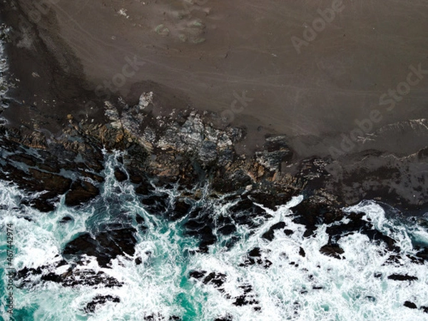Fototapeta Overhead view of a beach with black sand and the blue water of the sea crashing against the rocks on the shore.