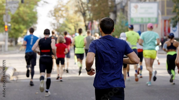 Fototapeta Man wearing blue t-shirt and earphones running on empty city road, blurred crowd of athletes running ahead. Contestants of sports event. Concept of healthy lifestyle