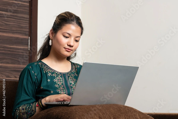 Fototapeta Young indian ethnic girl student freelancer working from home while sitting on the couch, looking at pc screen focused on thinking and solving online problem.