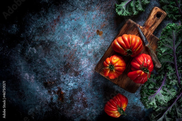 Fototapeta Flat lay of big fresh ripe tomatoes with kale leaves and wooden cutting board on the rustic dark blue background