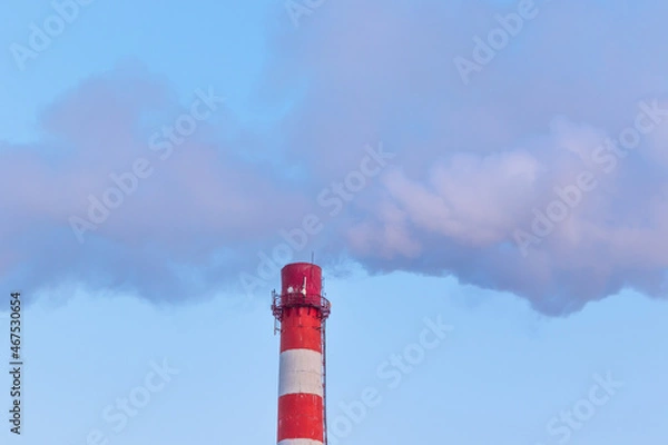 Fototapeta red and white chimney of a factory, from which gray smoke comes out against a blue sky on a sunny frosty winter day