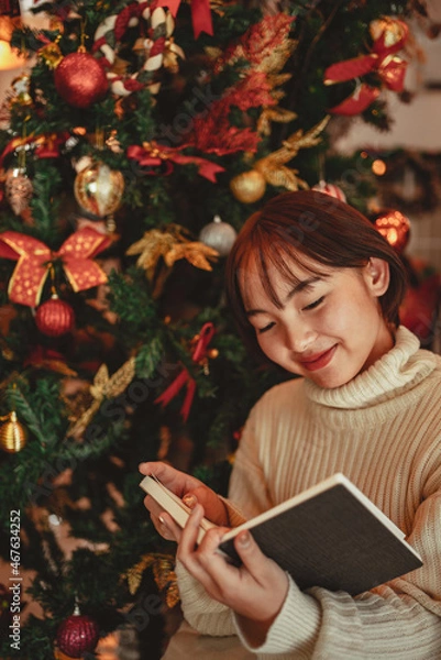Fototapeta a young Woman in front of Christmas tree reading a book at home with x-mas decoration.