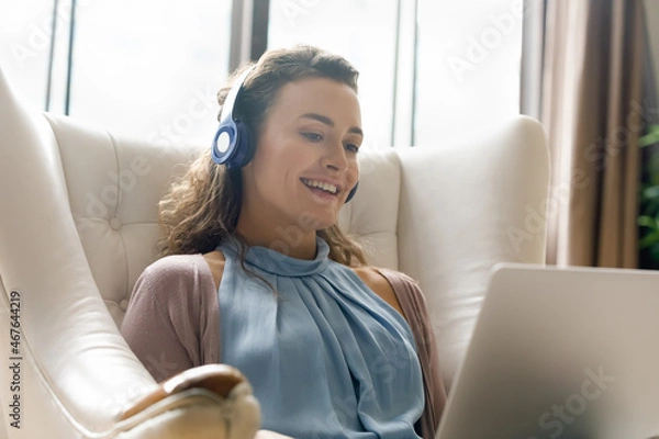 Fototapeta Happy young woman in wireless headphones looking at computer screen, watching funny movies or enjoying pleasant video call conversation, sitting in comfortable armchair, distant communication concept.