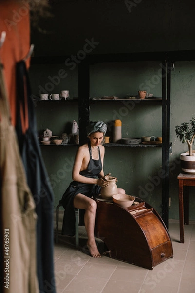 Fototapeta young sculptor, a girl with bandage on her head and in black apron, sculpts a clay product in pottery workshop near large window. Concept hobby sculpture