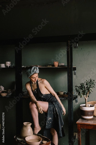 Fototapeta beautiful sculptor girl with bandage on her head and a black apron on her naked body is standing in pottery workshop near large window. concept is beauty and art.