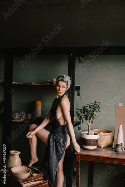Fototapeta beautiful sculptor girl with bandage on her head and a black apron on her naked body is standing in pottery workshop near large window. concept is beauty and art.