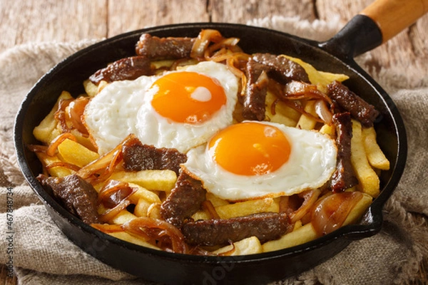 Fototapeta Traditional Chilean Chorrillana made of Fries with beef, caramelized onions, and fried eggs close up in the pan on the table. Horizontal