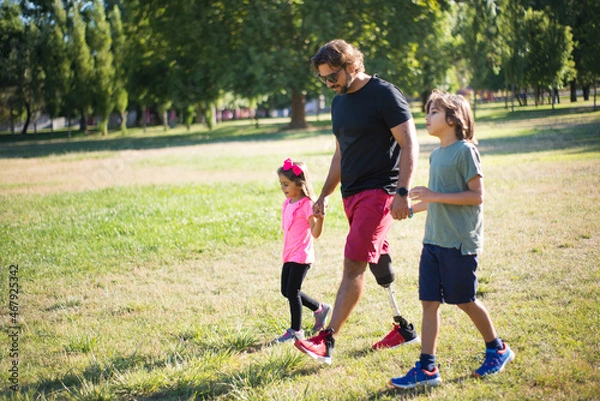 Fototapeta Side view of father with disability walking with children. Wavy-hairedman with mechanical leg having walk in park with little boy and girl. Disability, family, love concept