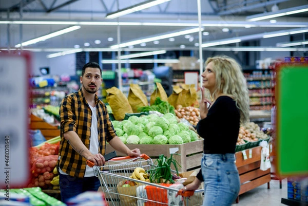 Fototapeta Couple in supermarket using smartphone with shopping cart while grocery shopping