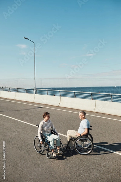 Fototapeta Vertical portrait of adult couple in wheelchairs chatting outdoors and enjoying city life, copy space