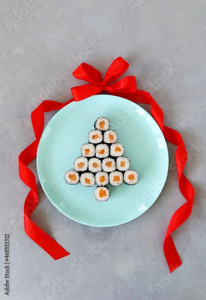 Fototapeta Christmas tree made of sushi on blue round plate with red bow and ribbon on a gray background. A creative idea for a Japanese restaurant. Vertical photo. Top view. Imitation of a Christmas ball.