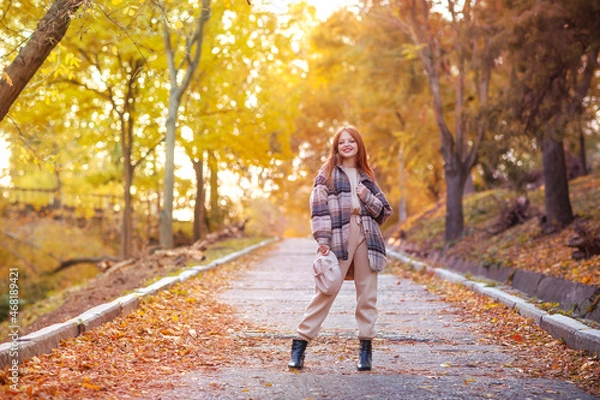 Fototapeta A young beautiful girl with long red hair stands right in the middle of the road going into the distance in an autumn park. Autumn outfit, fallen leaves, walk, good mood sunlight concept