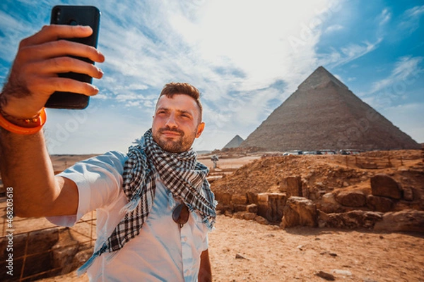 Fototapeta A young man, in front of the pyramid, raises his phone, takes a selfie with his fingers in front of the camera.