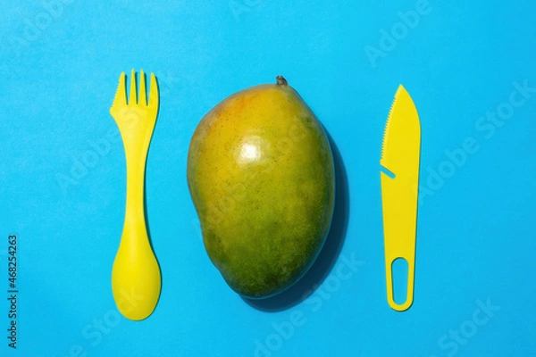 Fototapeta Whole mango fruit with a plastic knife and fork on a blue background. View from above