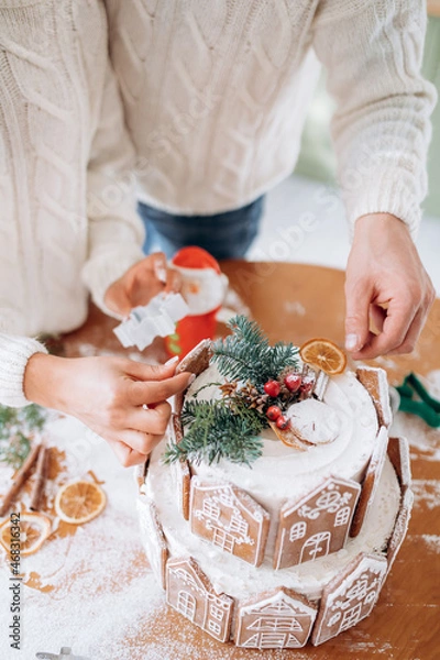 Fototapeta Gingerbread house and women's and man's hands in sweater, the concept of preparation for the Christmas holidays.