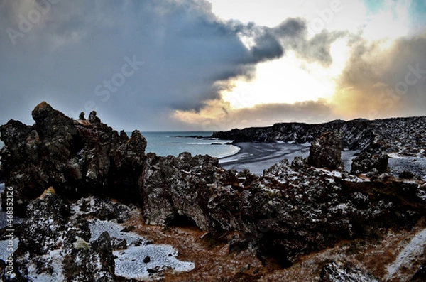 Obraz black basalt rocks covered with snow on the beach in the background sunbeams among the clouds Lóndrangar, Hellnar, Iceland