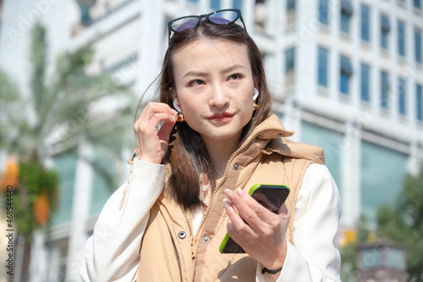Fototapeta Portrait of young Asian Chinese woman on the street wearing casual clothes and glasses on head has headphones in ears and phone listens to music and enjoys life dancing on street having fun catch vibe
