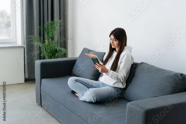 Fototapeta Indignant young woman sitting on couch in living room holding mobile phone having problems with digital device, feels angry and annoyed.