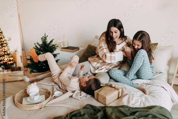 Fototapeta A happy mom with two daughters is resting, relaxing and having fun sitting on the bed in a cozy decorated bedroom during the Christmas holiday at home during the New Year vacations. Selective focus