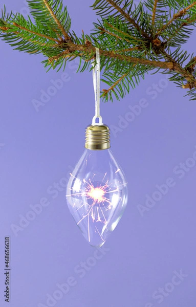Fototapeta Lightbulb with sparkler fireworks in it hanging from Christmas natural fir branch. Pastel purple background. Winter holidays and New Year idea.