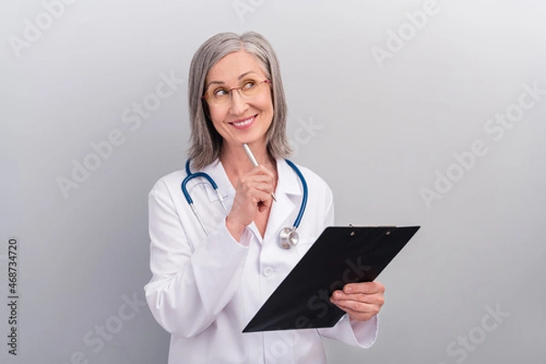Fototapeta Photo of happy dreamy lady doctor wear white coat spectacles smiling signing documents looking empty space isolated grey color background