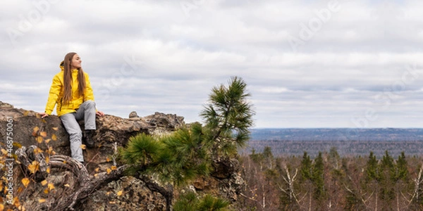 Fototapeta A smiling tourist, in a yellow jacket and gray pants, sits on the top of a mountain and admires the beauty of nature. Panoramic image.